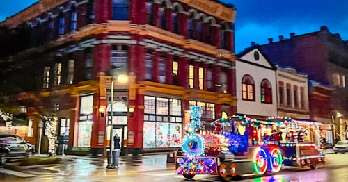 A festive parade float adorned with colorful lights moves past historic buildings on a rainy evening.