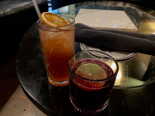 Two cocktails on a dark table: one tall with a slice of dried orange, the other short with a lime wedge.