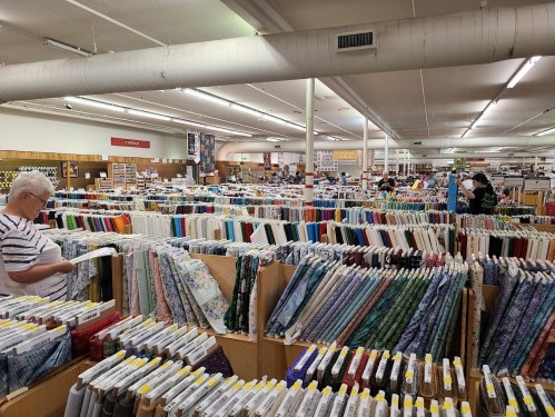 A spacious fabric store with rows of colorful fabric bolts and customers browsing among them.