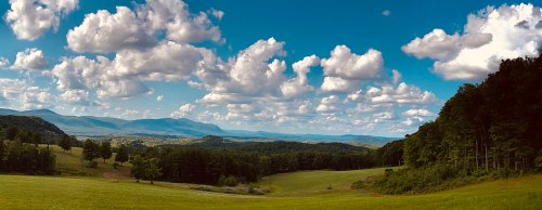 A panoramic view of rolling hills and lush greenery under a bright blue sky with fluffy white clouds.