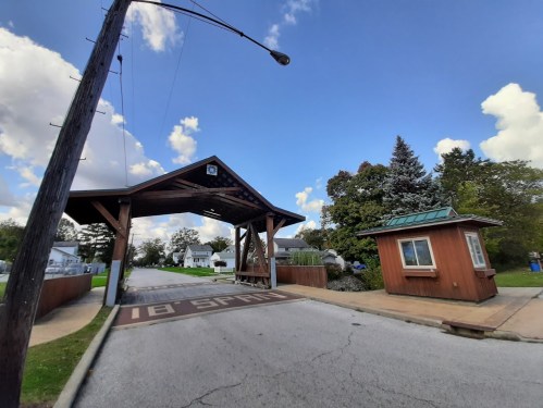 A wooden entrance structure with a small guard booth beside a road under a blue sky with scattered clouds.