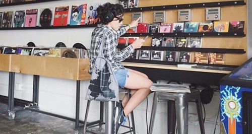 A person sits on a stool in a record store, browsing through vinyl records while wearing headphones.