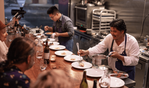 A chef serves dishes at a bustling restaurant bar, with diners enjoying the culinary experience.