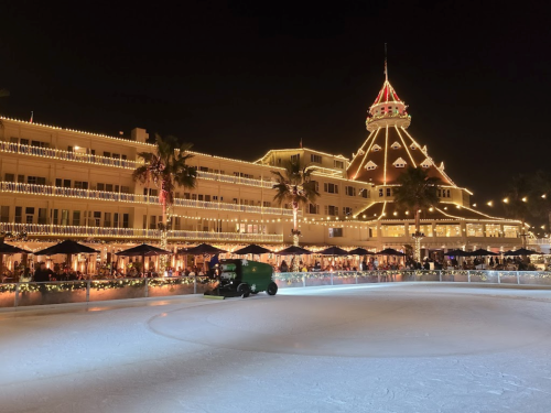 A nighttime view of an ice rink with a Zamboni, surrounded by a beautifully lit building and festive decorations.