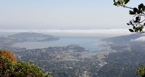 A scenic view of a bay with hills, a bridge, and a coastal town under a clear sky with some clouds.