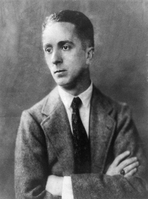 A black-and-white portrait of a young man in a suit, looking thoughtfully to the side with arms crossed.