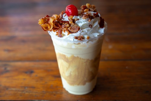 A creamy milkshake topped with whipped cream, crispy bacon bits, and a cherry, served in a clear cup.