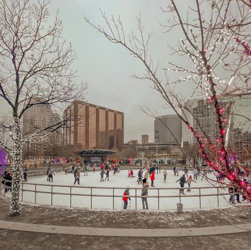 A bustling ice skating rink surrounded by trees adorned with colorful lights and city buildings in the background.