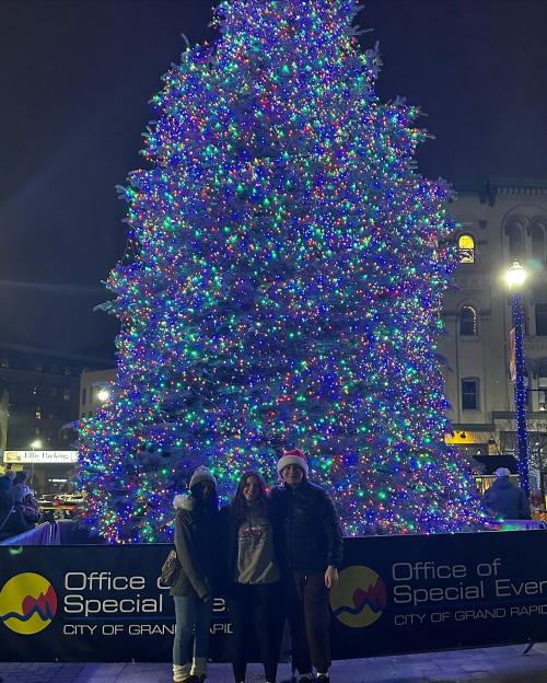 Three people stand in front of a large, brightly lit Christmas tree at night, with festive decorations and a city backdrop.