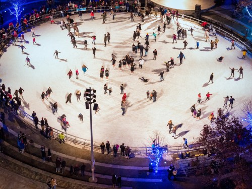Aerial view of a busy ice skating rink with many skaters and spectators, surrounded by festive lights and trees.