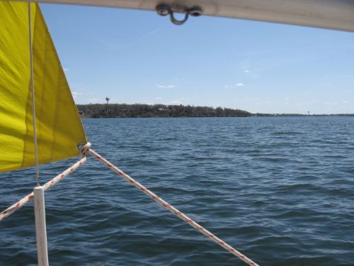 View from a sailboat with a yellow sail, showing calm water and a distant shoreline under a clear blue sky.