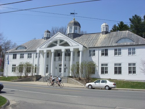 A white, historic building with a large front porch and dome, surrounded by trees and a road, with cyclists passing by.