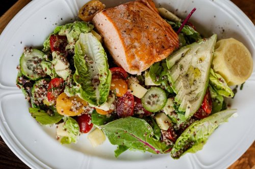 A plate of salad with mixed greens, cherry tomatoes, cucumbers, quinoa, and a piece of grilled salmon on top.
