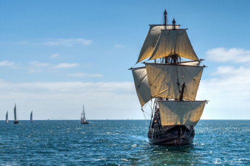 A tall ship with billowing sails navigates a calm sea under a clear blue sky, with smaller boats in the background.