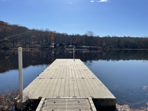 A wooden dock extends into a calm lake, surrounded by trees with autumn foliage under a clear blue sky.