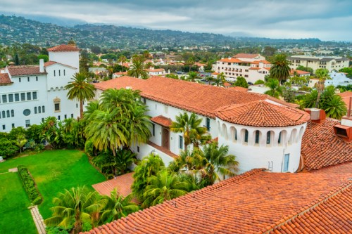Aerial view of a Spanish-style building with red tile roofs, surrounded by lush greenery and hills in the background.
