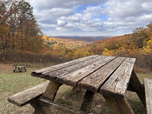 A weathered wooden picnic table overlooks a colorful autumn landscape with rolling hills and cloudy skies.