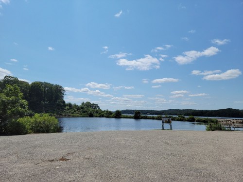 A serene landscape featuring a calm lake surrounded by greenery under a clear blue sky with scattered clouds.