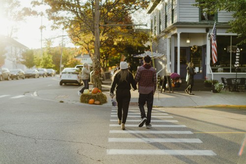 A couple walks hand in hand across a crosswalk, surrounded by autumn scenery and a quaint town.