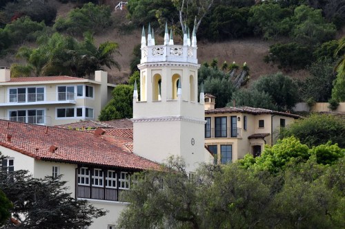 A white tower with decorative features rises above a cluster of homes surrounded by greenery and hills.