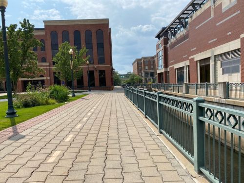 A paved walkway beside a canal, flanked by modern brick buildings and greenery under a partly cloudy sky.