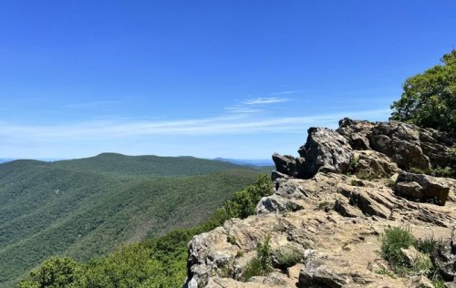 A rocky outcrop overlooks a lush green valley under a clear blue sky.