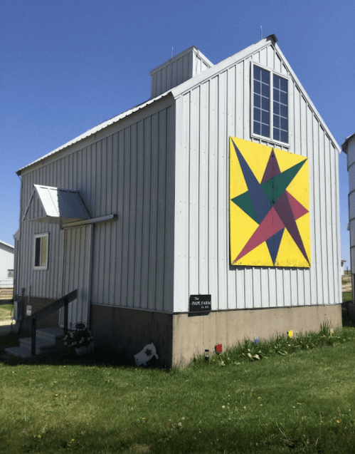 A silver barn with a colorful star quilt pattern on the side, set against a clear blue sky and green grass.
