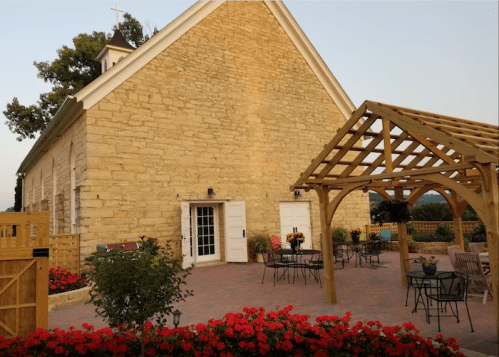 A stone building with a wooden pergola and flower beds, featuring outdoor seating in a serene setting.