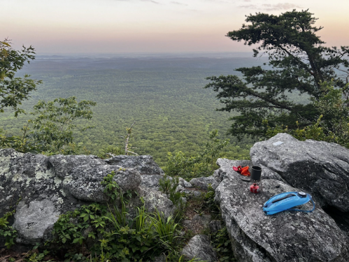 A scenic view from a rocky ledge overlooking a lush green forest at sunset, with camping gear nearby.