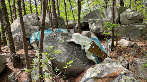 A partially buried, weathered piece of metal debris among large rocks and trees in a forested area.