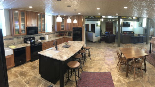 A spacious kitchen with wooden cabinets, a large island, and a dining table, featuring stone flooring and modern appliances.