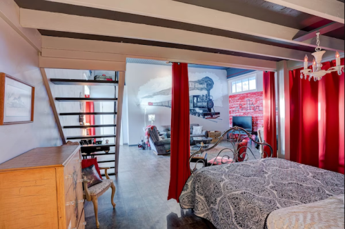 Cozy interior featuring a bedroom with a patterned bedspread, stairs, and a living area with a mural and red curtains.