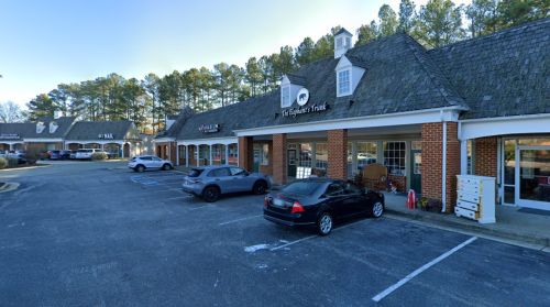 A row of shops in a plaza with parked cars and trees in the background on a clear day.