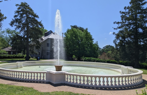 A circular fountain with a tall spray, surrounded by greenery and a historic building in the background.