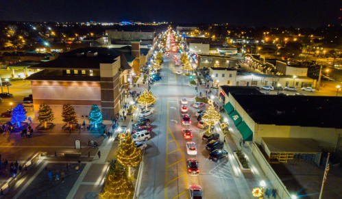 Aerial view of a festive street at night, lined with holiday lights, decorated trees, and parked cars.