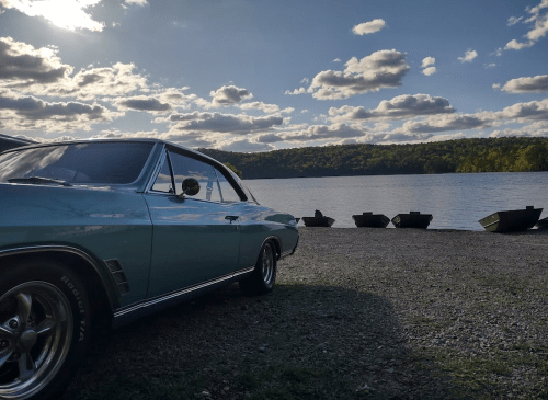 A vintage blue car parked by a serene lake, with boats lined up and fluffy clouds in a bright sky.