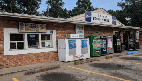 Exterior of Nelson Food Market with vending machines and a sign, surrounded by trees and parked cars.