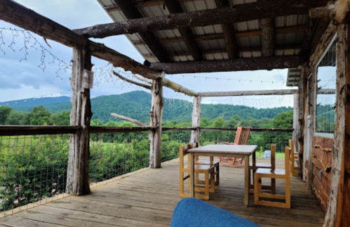 A rustic porch with wooden furniture overlooks a green landscape and mountains under a cloudy sky.