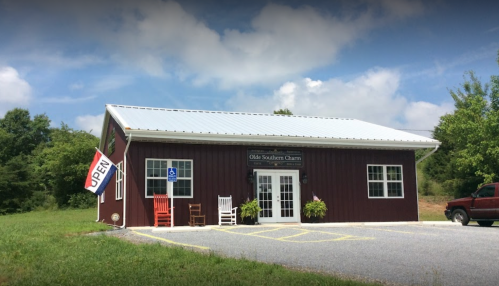 A red barn-style shop with a metal roof, featuring rocking chairs outside and an "OPEN" sign. Green grass and trees in the background.