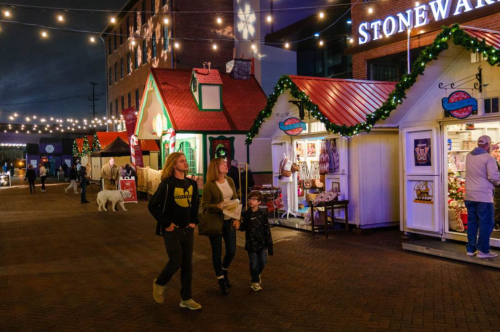 People walking past festive market stalls decorated with lights and greenery in an evening setting.