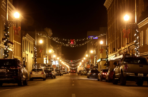 A festive street at night, lined with decorated buildings and cars, featuring holiday lights and ornaments.