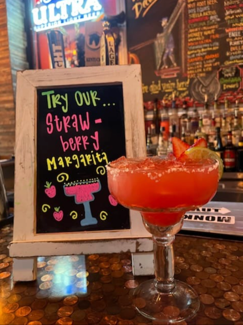 A vibrant strawberry margarita in a glass, next to a colorful sign promoting the drink.