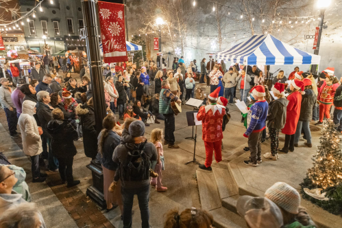 A festive crowd gathers around performers in Santa hats at a holiday market, with lights and tents in the background.