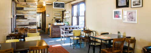 Cozy café interior with wooden tables, chairs, and a counter in the background, featuring warm lighting and decor.