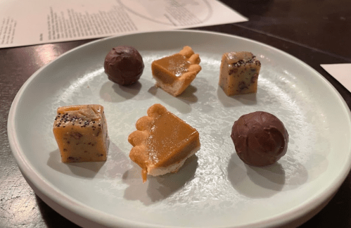 A plate with six small desserts: two chocolate truffles, two caramel squares, and two pastry bites.