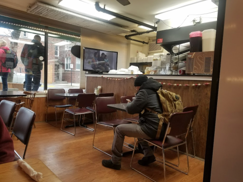 A person with a backpack sits alone at a table in a cozy diner, with a view of people outside through the window.