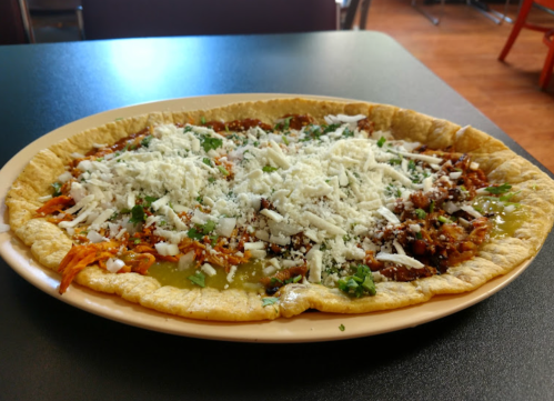 A large plate of tortilla topped with shredded meat, cheese, and fresh herbs, served in a casual dining setting.
