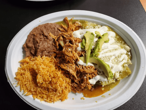A plate of shredded chicken with green sauce, rice, refried beans, and avocado slices, drizzled with sour cream.