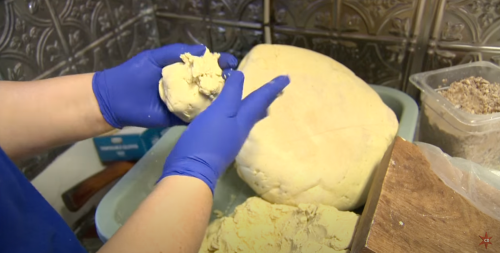 A person in blue gloves is shaping dough in a kitchen, with a large ball of dough and other ingredients nearby.