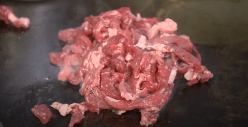 Sizzling strips of raw beef on a hot cooking surface, with steam rising around them.